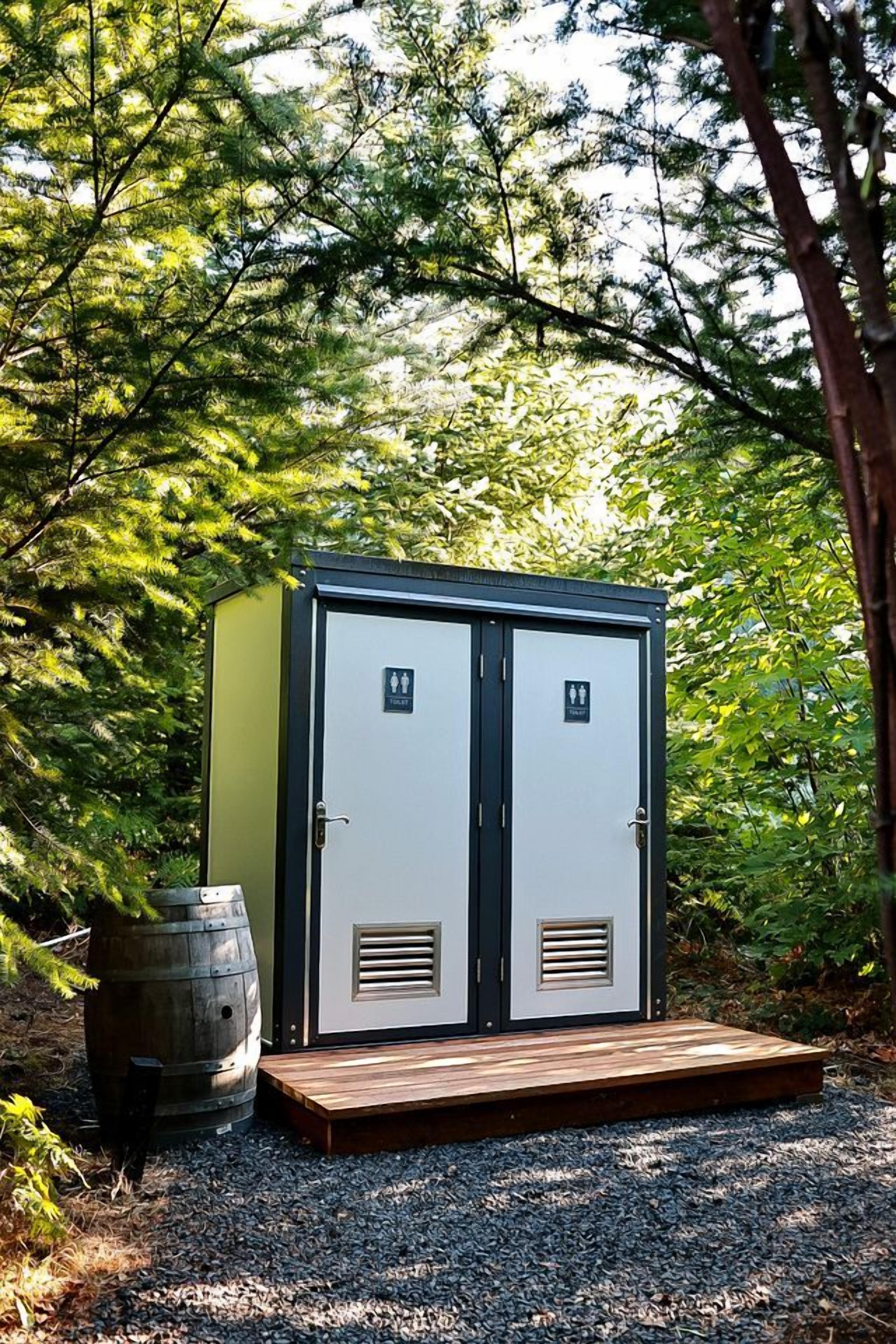 Un baño portátil con dos puertas, cada una con un símbolo de género, sobre una plataforma de madera, con un barril cercano, rodeado de árboles.