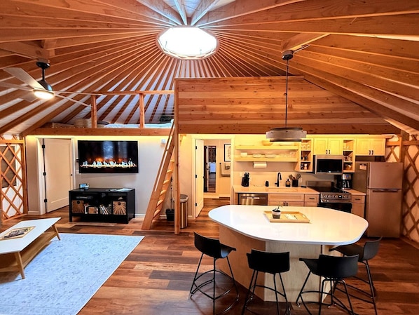 A loft-style interior with a central island, wooden beams, a TV, kitchen appliances, and a staircase. - Modern Luxury Yurt with Firepit Table & Skylit Dome near Carson, Washington (Carson)