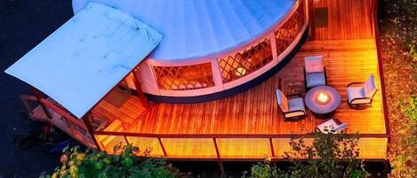 Une terrasse circulaire en bois, dotée d’un foyer et d’un espace salon, entourée d’une végétation luxuriante.