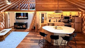 A loft-style interior with a central island, wooden beams, a TV, kitchen appliances, and a staircase. - Modern Luxury Yurt with Firepit Table & Skylit Dome near Carson, Washington (Carson)