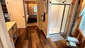 A compact, wood-paneled bathroom with a shower, a toilet, and a small bedside table. - Modern Luxury Yurt with Firepit Table & Skylit Dome near Carson, Washington (Carson)
