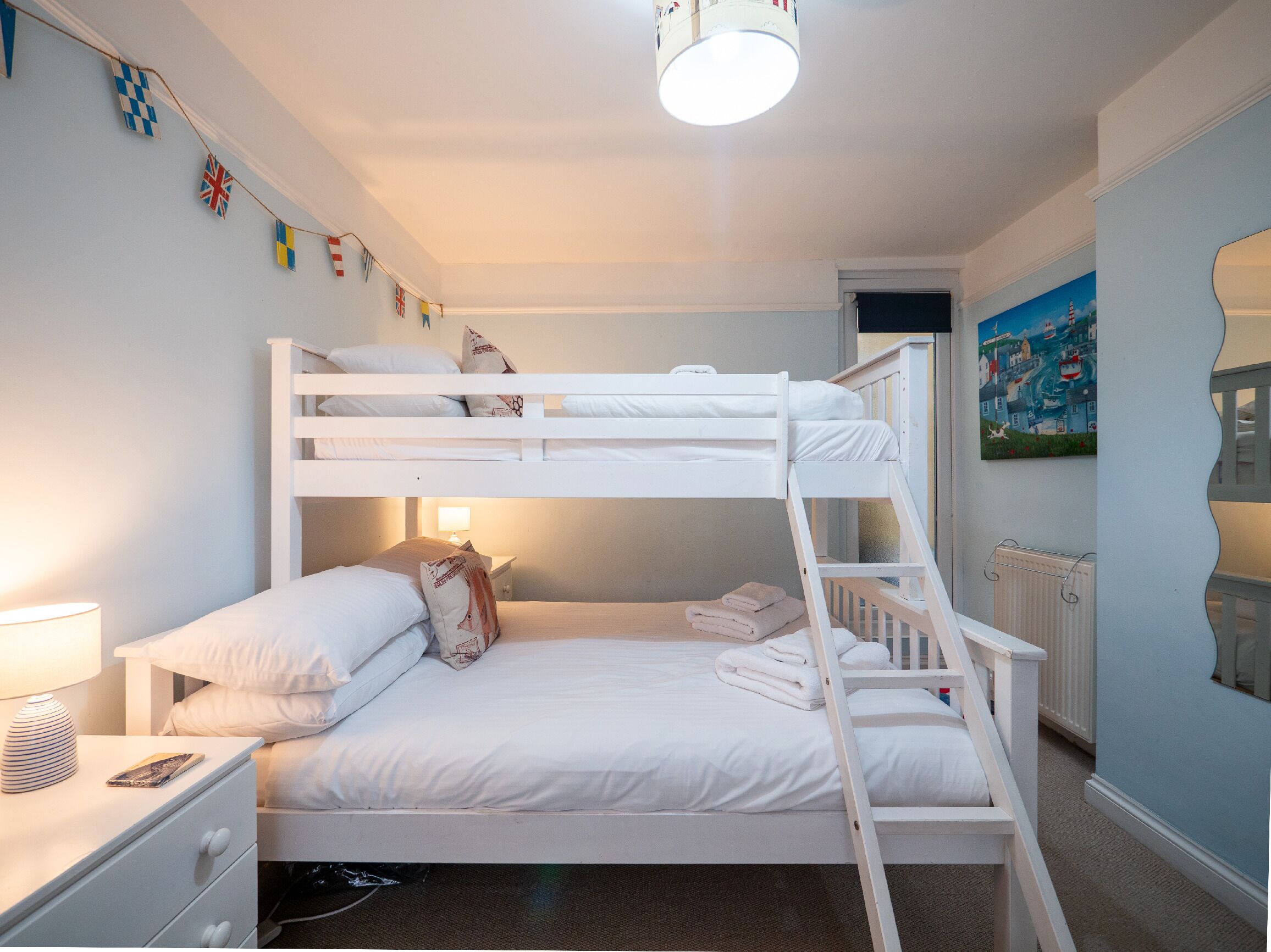 A bunk bed room with white bedding, a nightstand with a lamp, and a painting on the wall.