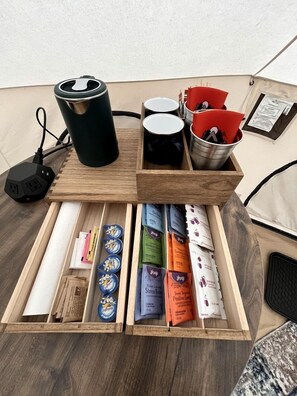 A wooden table with a black power adapter, a black mug, red napkins, and a drawer containing various packaged items.