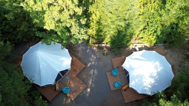 Eine Luftaufnahme eines Campingplatzes mit zwei großen weißen Sonnenschirmen, Holzterrasse und umliegender Bepflanzung.