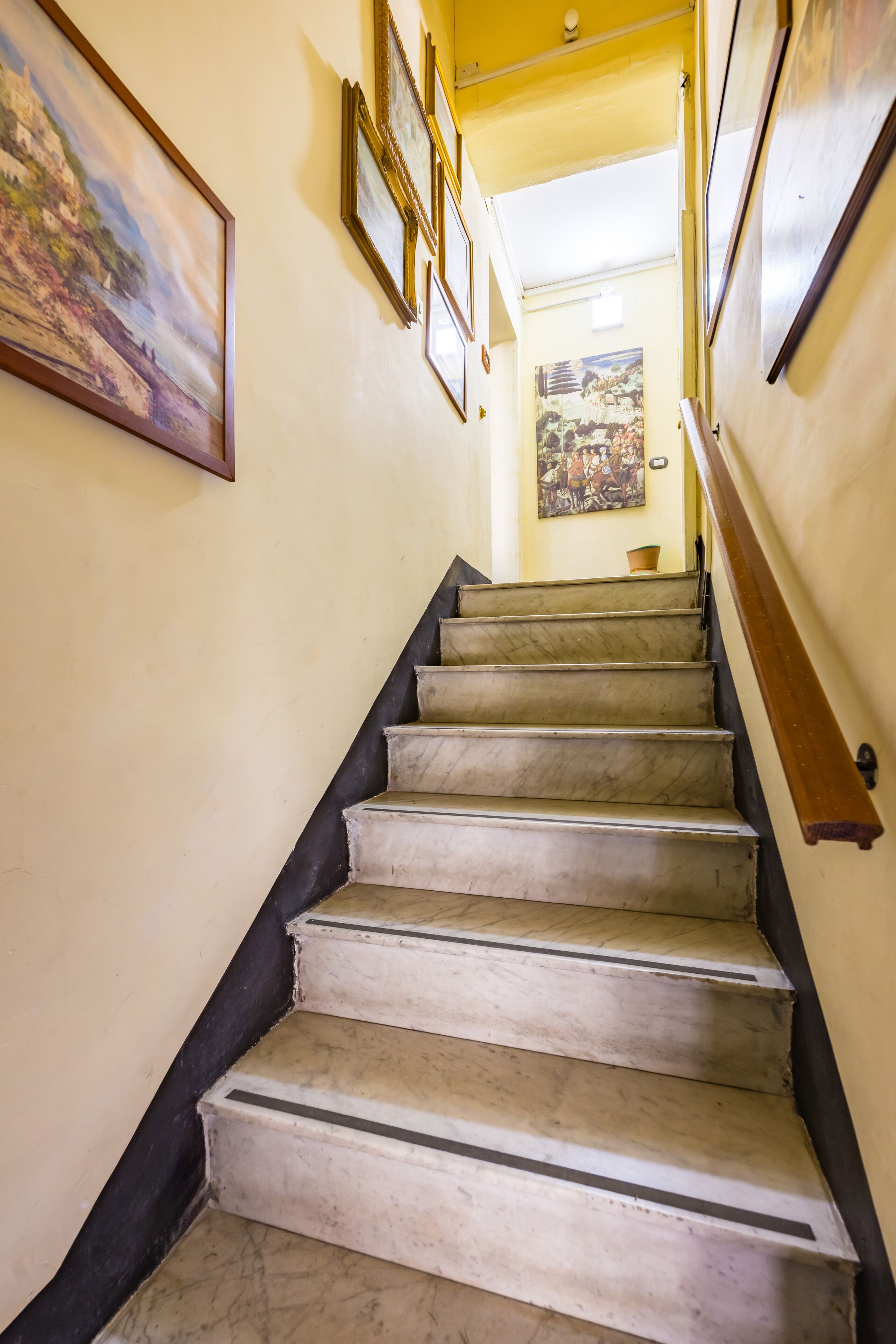 Un escalier en marbre avec des rampes noires, menant à une pièce aux murs jaunes et ornée d’œuvres d’art encadrées.