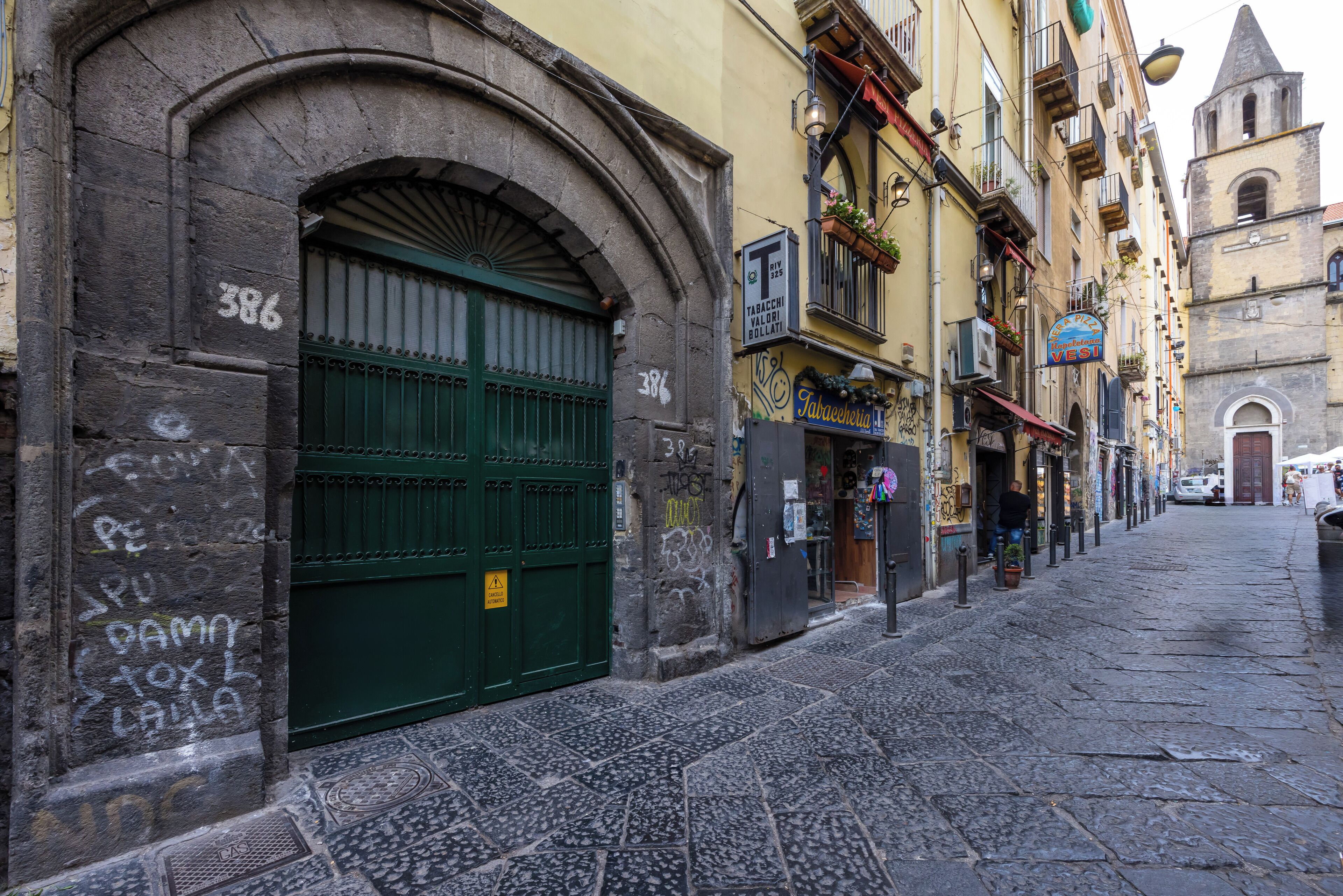 Uma rua estreita com um portão verde, um edifício com a placa 'Tabacchi e Follati', e uma torre de igreja ao fundo.