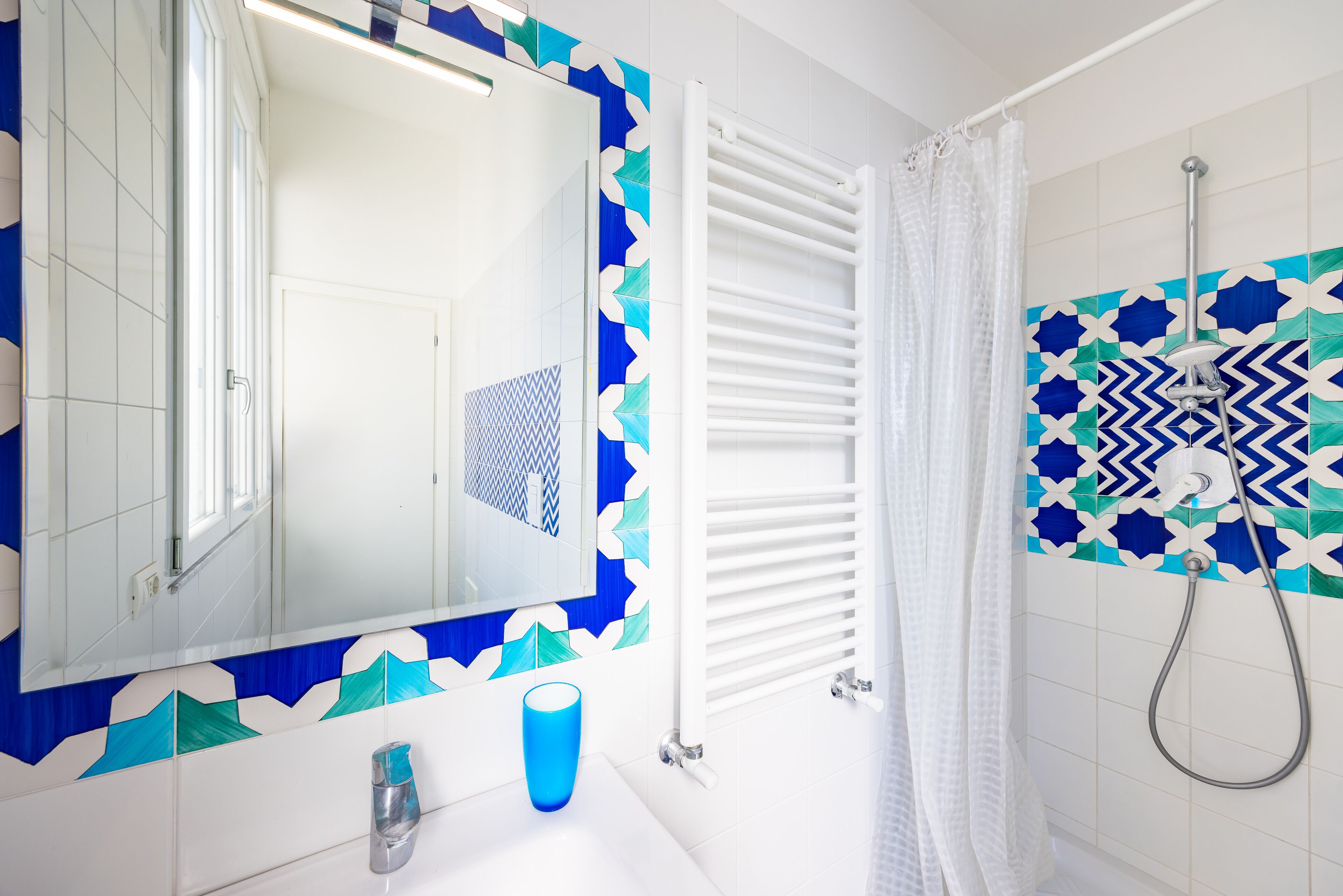Un baño con un lavamanos blanco, un espejo, una taza azul, una ducha con pared de azulejos estampados y un toallero blanco.