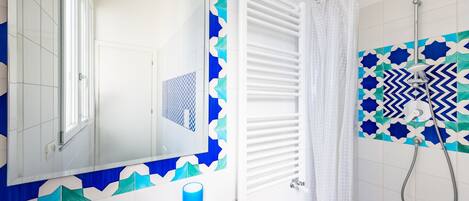 Un baño con un lavamanos blanco, un espejo, una taza azul, una ducha con pared de azulejos estampados y un toallero blanco.