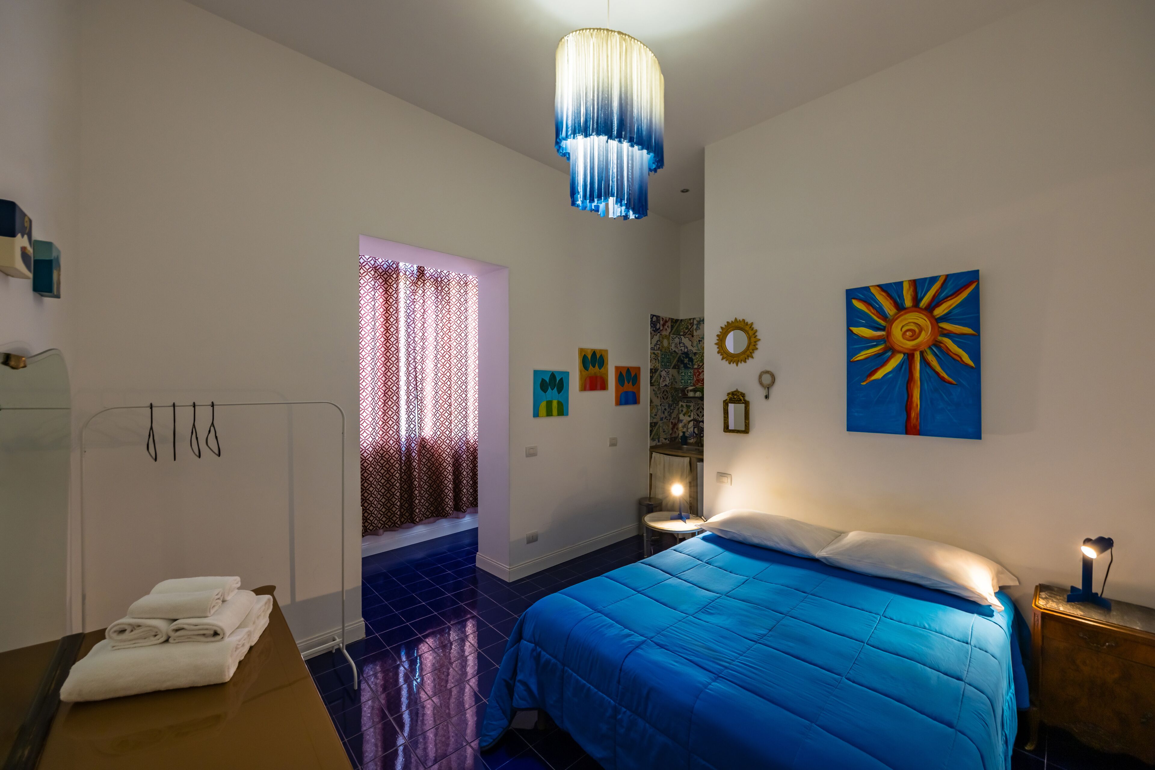 A bedroom with a bed, a bedside table, a nightstand, a hanging light fixture, and a window with patterned curtains.