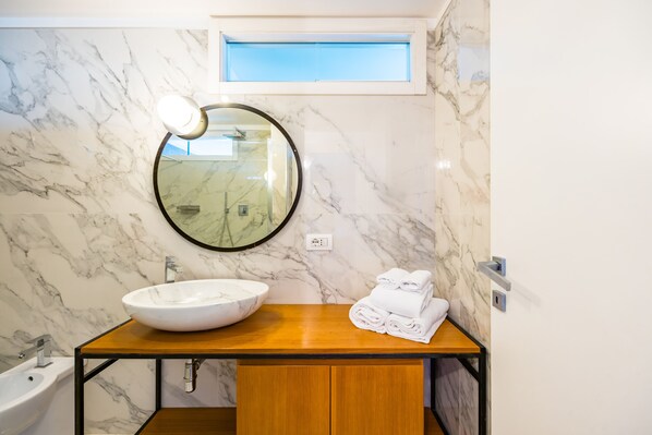 Une salle de bain avec un mur en marbre, un miroir rond, un meuble-lavabo en bois, un lavabo et une fenêtre.