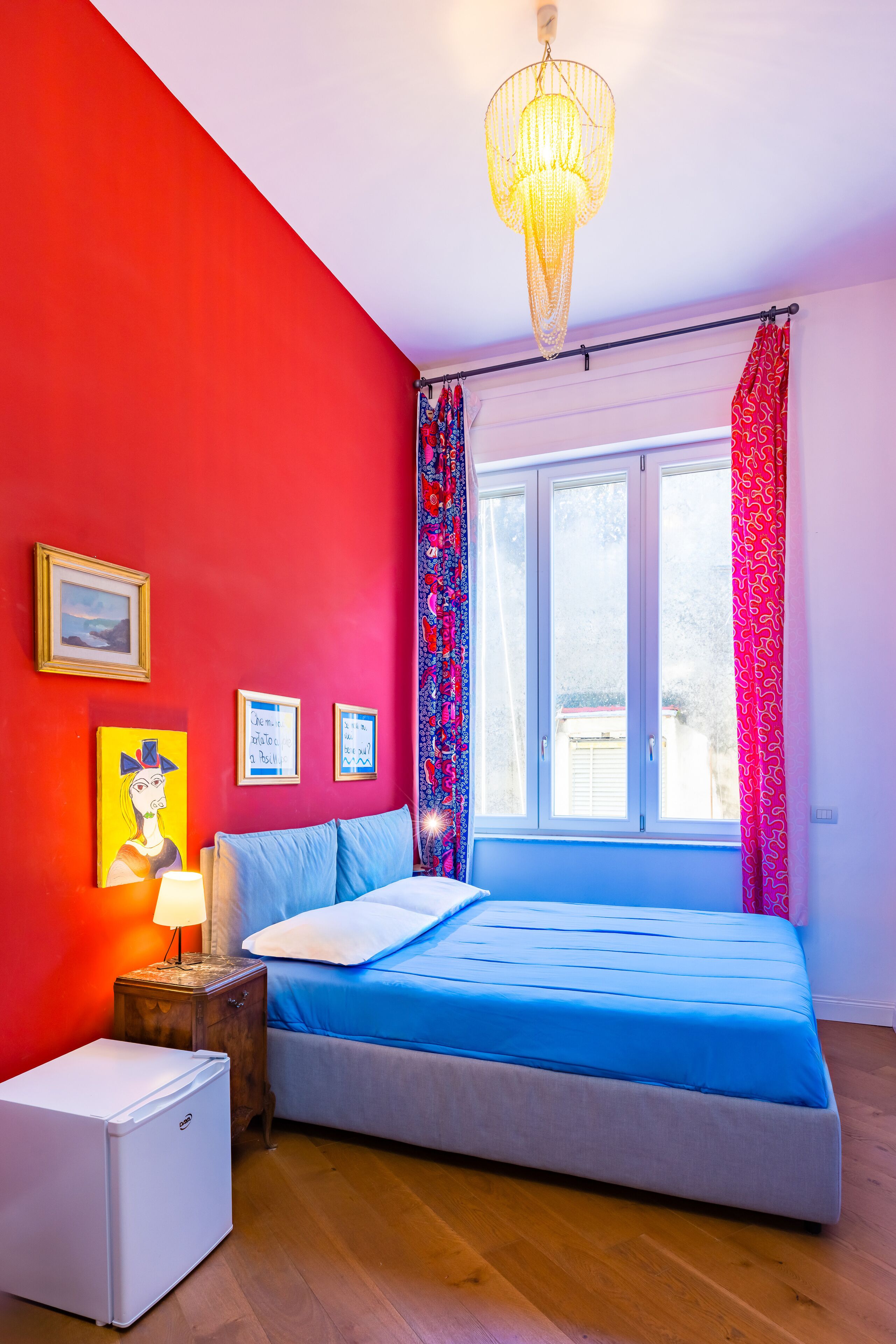 A bedroom with a bed, a small bedside table, a lamp, a refrigerator, a window with curtains, and wall art.
