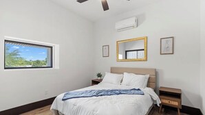Un dormitorio con cama, ventana, ventilador de techo, aire acondicionado instalado en la pared y cuadros enmarcados.
