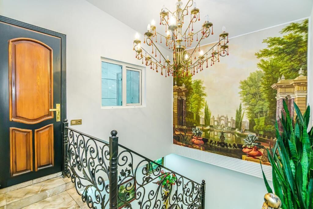 Un couloir avec une porte en bois, une fenêtre, une rampe en métal décorative et une fresque représentant un jardin.