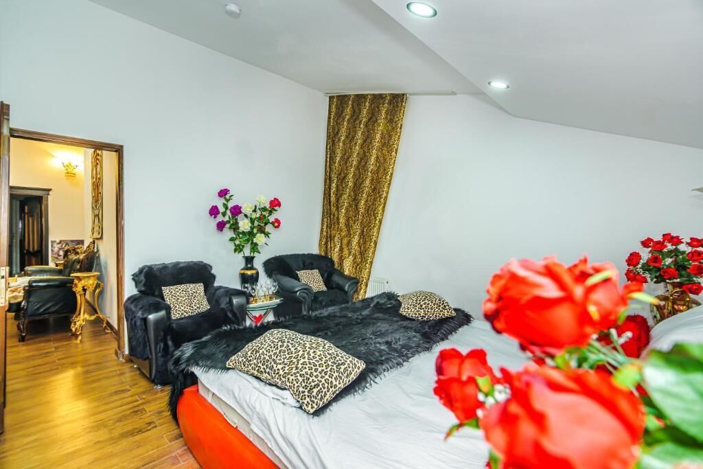 A modern bedroom with a large bed, two black armchairs, a gold curtain, and a vase of flowers.