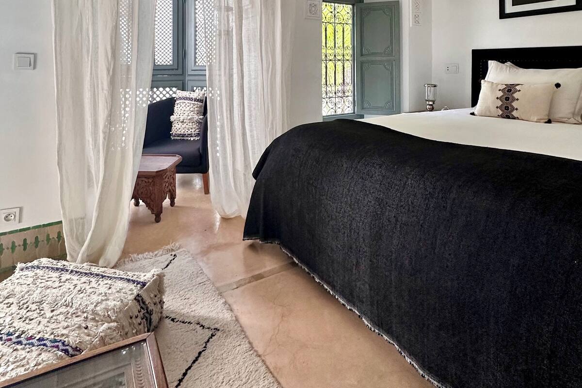 Une chambre avec un grand lit, un canapé et une petite table. Il y a une fenêtre avec des rideaux, une porte et une lampe fixée au mur.