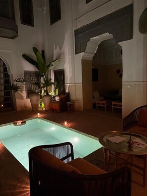 Un espace piscine agrémenté d’une chaise et d’une table, entouré de plantes en pot et éclairé par une lumière chaleureuse.