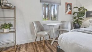 A bedroom with a bed, a desk, two chairs, a window with blinds, a plant, and a painting on the wall.