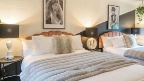A hotel room with two beds, each with a headboard, white bedding, and a fluffy throw blanket. There are framed pictures on the wall, a bedside table with a lamp, and a small plant.