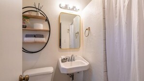 A bathroom with a toilet, sink, and mirror. There is a towel rack with towels and a toilet paper holder. A shower curtain is partially visible. - B. Abstract: Professional Design + King Bed! (Bandon)