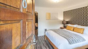 A hotel room with a wooden wardrobe, a bed with white bedding and a grey throw, a nightstand with a lamp, and a patterned headboard.