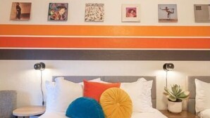 Un lit avec des coussins colorés, une table de chevet avec une lampe et un mur orné de tableaux encadrés.