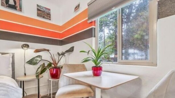 Une pièce de taille modeste, avec un lit blanc, une petite table sur laquelle se trouve un pot rouge, et une fenêtre donnant sur des arbres.