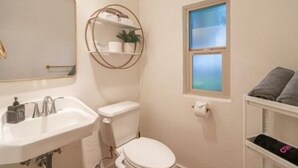 Een kleine badkamer met een witte wastafel, toilet en een ronde spiegel. Er is een raam met uitzicht op de lucht en een plankje met handdoeken en toiletartikelen.