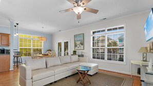 A spacious living room with a beige sectional sofa, a small round coffee table, and a ceiling fan. There is a dining area with a table and chairs, and a kitchen area with wooden cabinets and a countertop. - Surfside Retreat w/ Pool & Elevator Access (Surfside Beach)