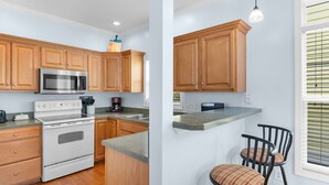 A kitchen with wooden cabinets, a stainless steel microwave, and a built-in oven. There are two bar stools and a window with blinds. - Surfside Retreat w/ Pool & Elevator Access (Surfside Beach)