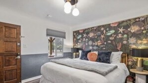 A bedroom with a large bed, a wooden door, a window, and a wall with a floral mural. - b. under the sea #2 King bedroom (Bandon)