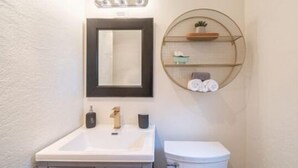 A bathroom with a white sink, a mirror, a toilet, and a round shelf with toiletries. - b. under the sea #2 King bedroom (Bandon)