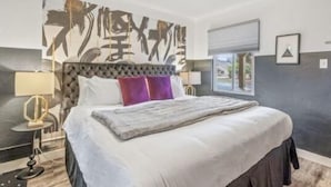 A modern bedroom with a tufted headboard, a large abstract painting on the wall, a bed with white and purple pillows, and a window with blinds. - b.fresh #6 King bedroom (Bandon)