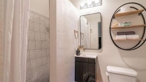 A bathroom with a toilet, sink, and mirror. There are shelves above the mirror and a shower curtain partially visible. - b.fresh #6 King bedroom (Bandon)