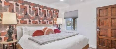 Ein Schlafzimmer mit Bett, Nachttisch, Lampe, einer Wand mit geometrischem Muster und einer Holztür.