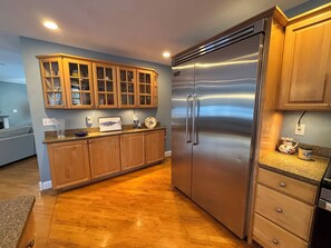 A modern kitchen with wooden cabinets, a stainless steel refrigerator, and a granite countertop. - Half-Acre Coastal Retreat- Hot tub and walk to town! (Ogunquit)