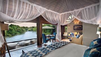 A bedroom with a large bed, a sitting area with a table and chairs, and a view of a river.