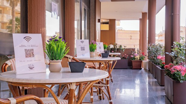 Un pasillo de hotel con mesas redondas, sillas de mimbre, plantas en macetas y códigos QR en los menús.
