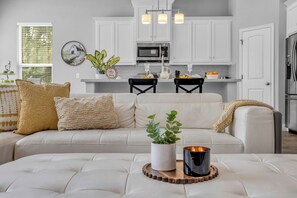 A modern living room with a white sofa, decorative pillows, a coffee table with a candle and a plant, and a kitchen in the background with white cabinets and a microwave. - 5 BR Houston Retreat - Private Loft + Huge Deck - Sleeps 12 (Houston)