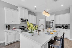 A modern kitchen with white cabinetry, stainless steel appliances, a central island with a plant, and a hanging light fixture.