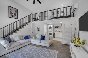 A modern living room with a grey sectional sofa, a staircase, and a kitchen area. - 5 BR Houston Retreat - Private Loft + Huge Deck - Sleeps 12 (Houston)
