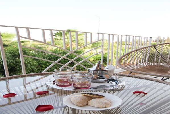 A round glass table with a tray holding glasses, a teapot, and cookies, set on a balcony with a view of greenery.