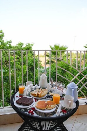 A breakfast table with various food items and drinks on a balcony with a view of greenery.