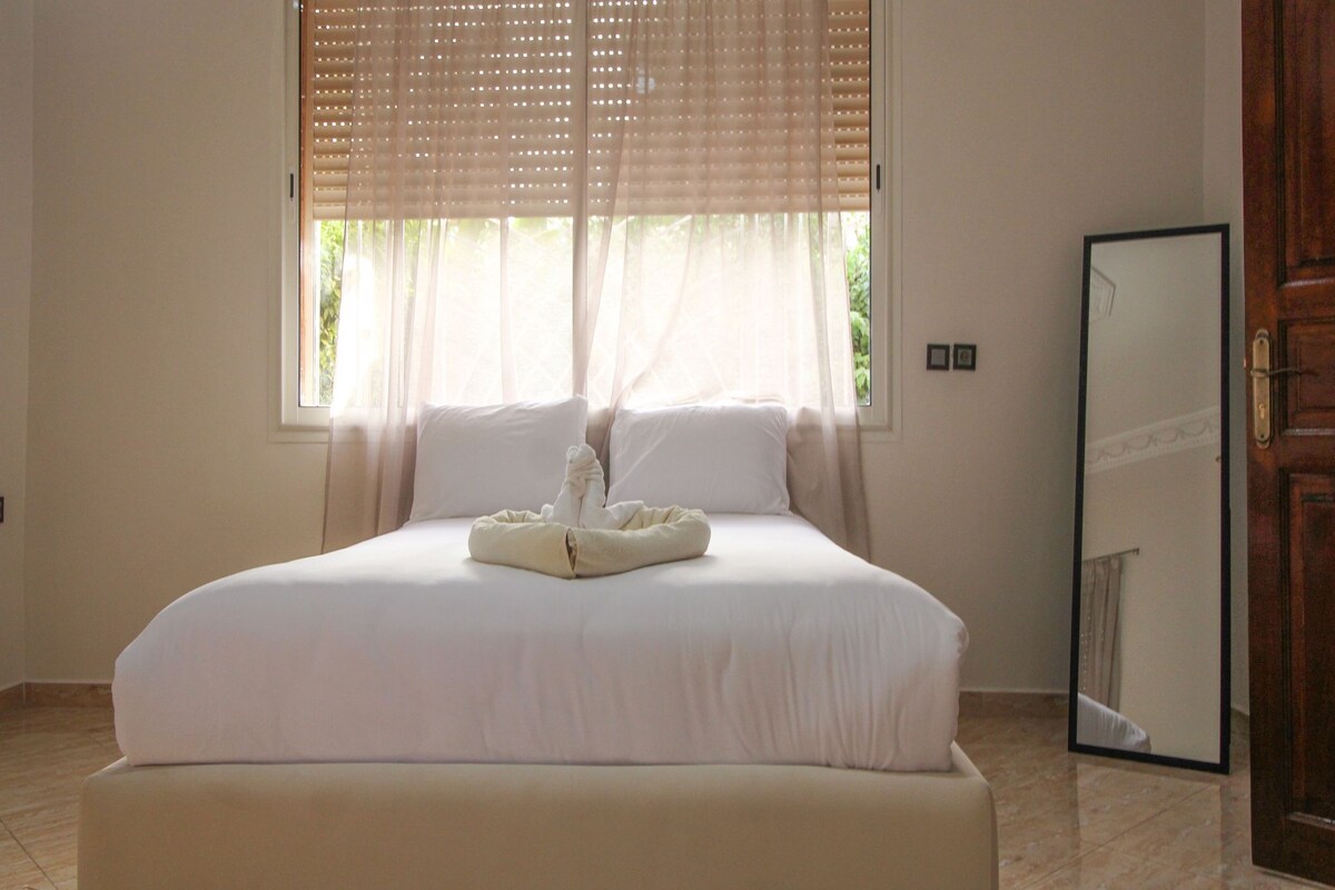 Una cama bien tendida con sábanas blancas y una toalla doblada, una ventana con cortinas translúcidas y una puerta de madera a la derecha.