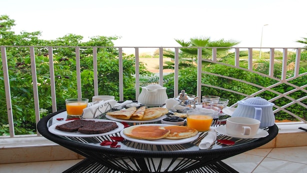 Et morgenbord med tallerkener med mad, kopper og et thestel på en balkon med udsigt til grønne områder.