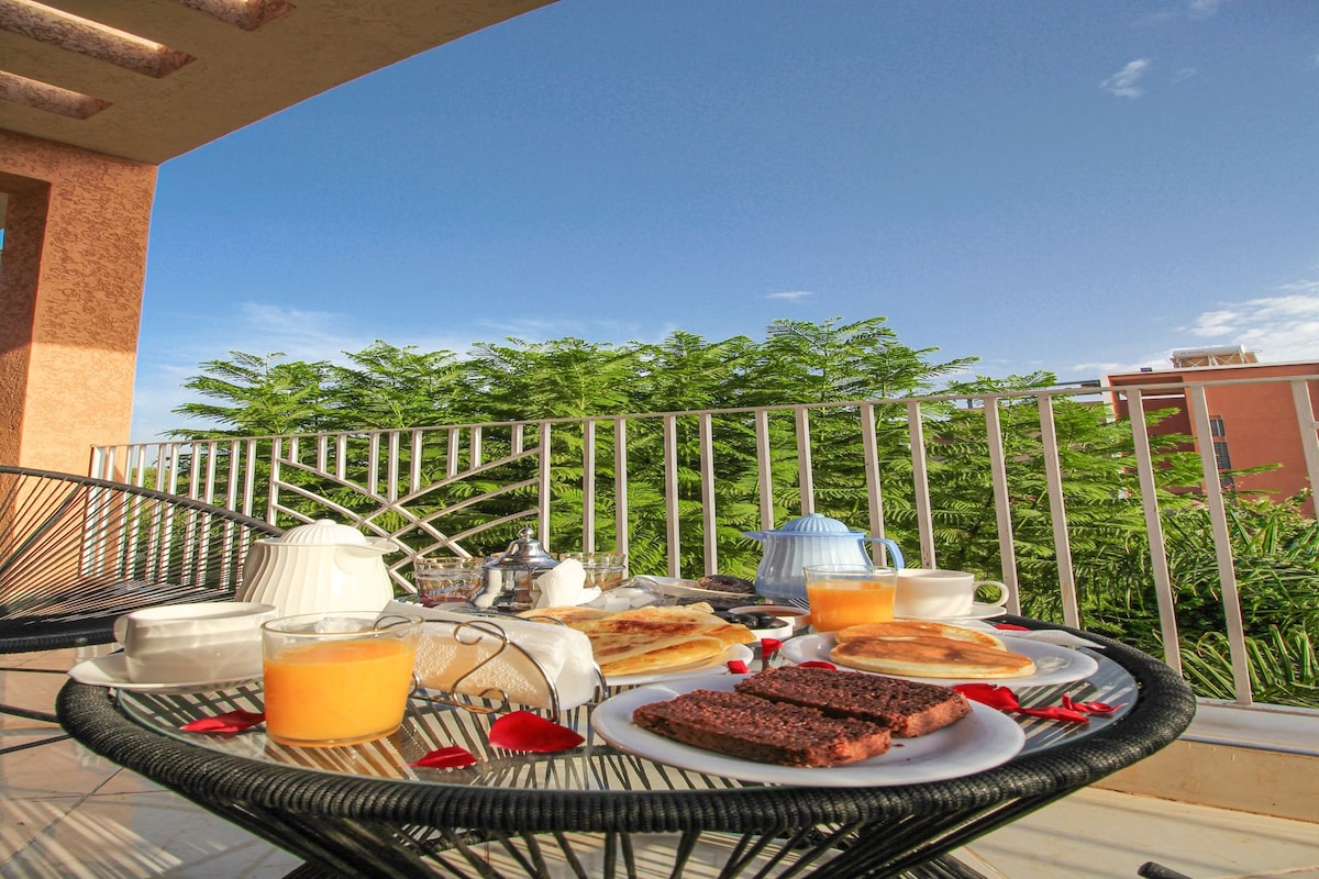 A dining table with breakfast items on a balcony with a view of greenery and buildings.