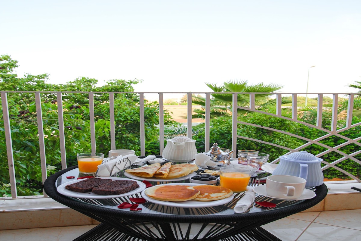 A breakfast table with plates of food, cups, and a teapot on a balcony with a view of greenery.