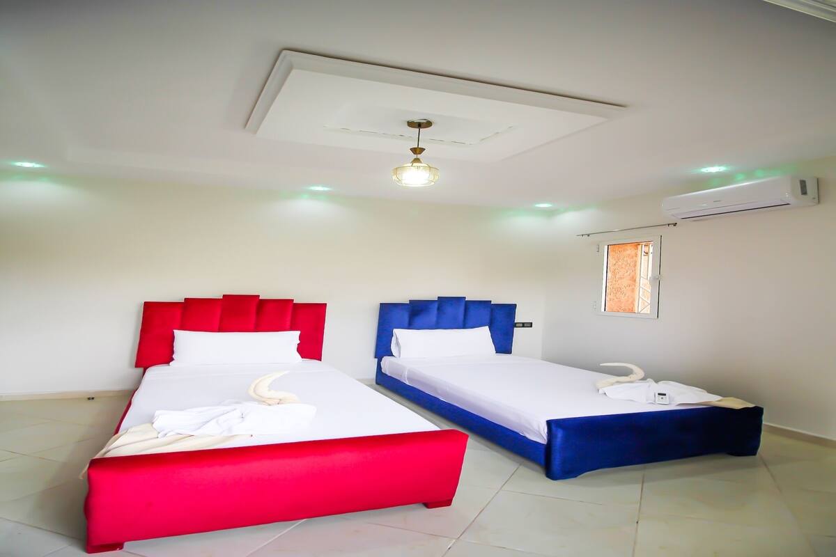 A hotel room with two beds, one with red and white bedding and the other with blue and white bedding. There is a ceiling fan, an air conditioning unit, and a window with a view.