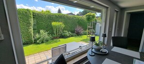 A room with a large window overlooking a well-maintained garden, a small table with a lamp, and a chair.