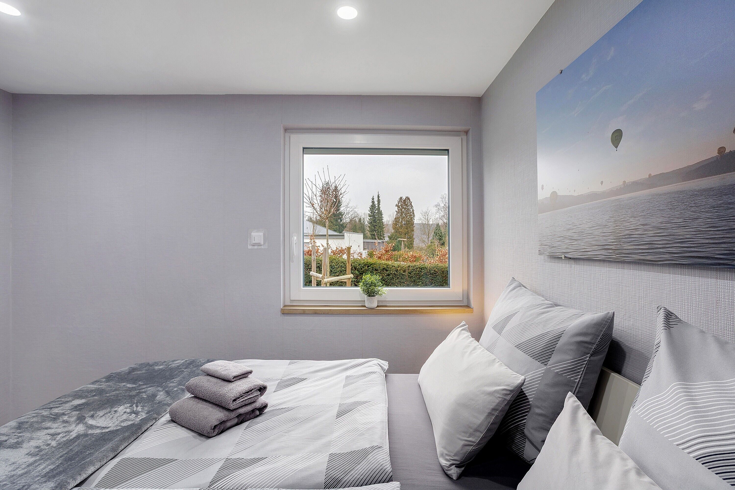 A bedroom with a bed, pillows, a window with a view, and a painting on the wall.
