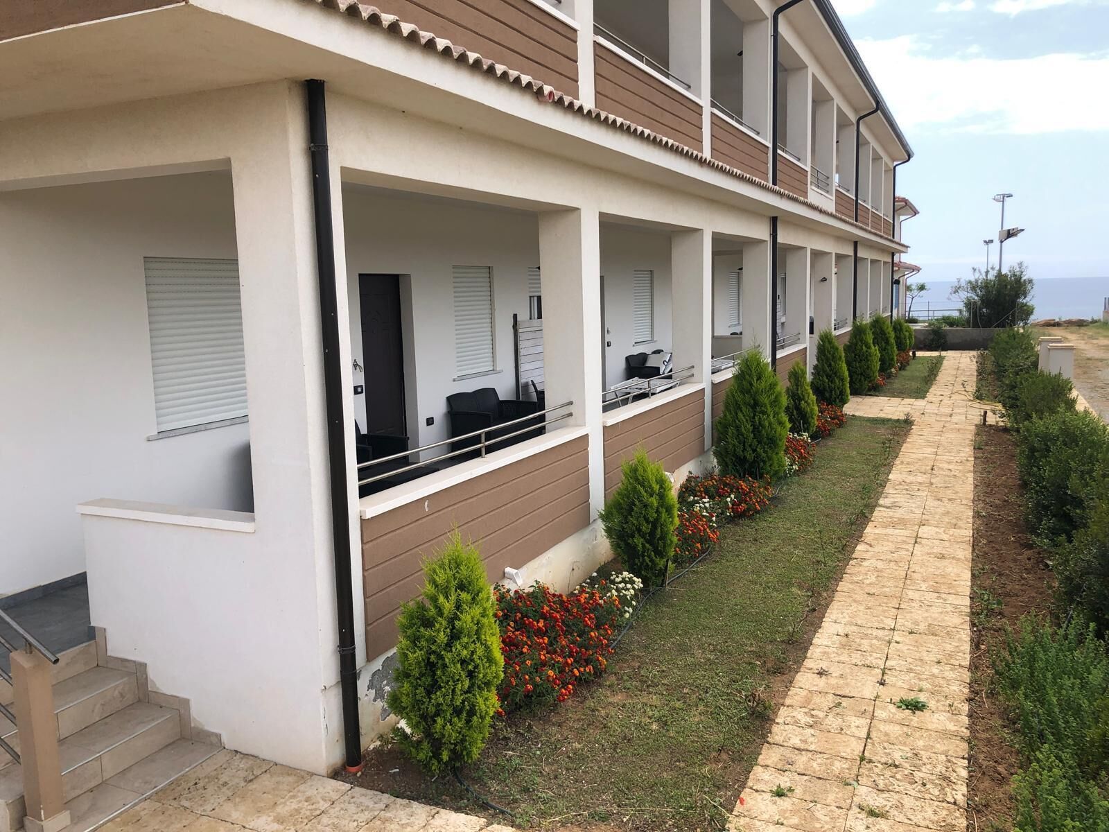 A modern hotel with a covered outdoor seating area, a well-maintained garden, and a clear view of the sea.